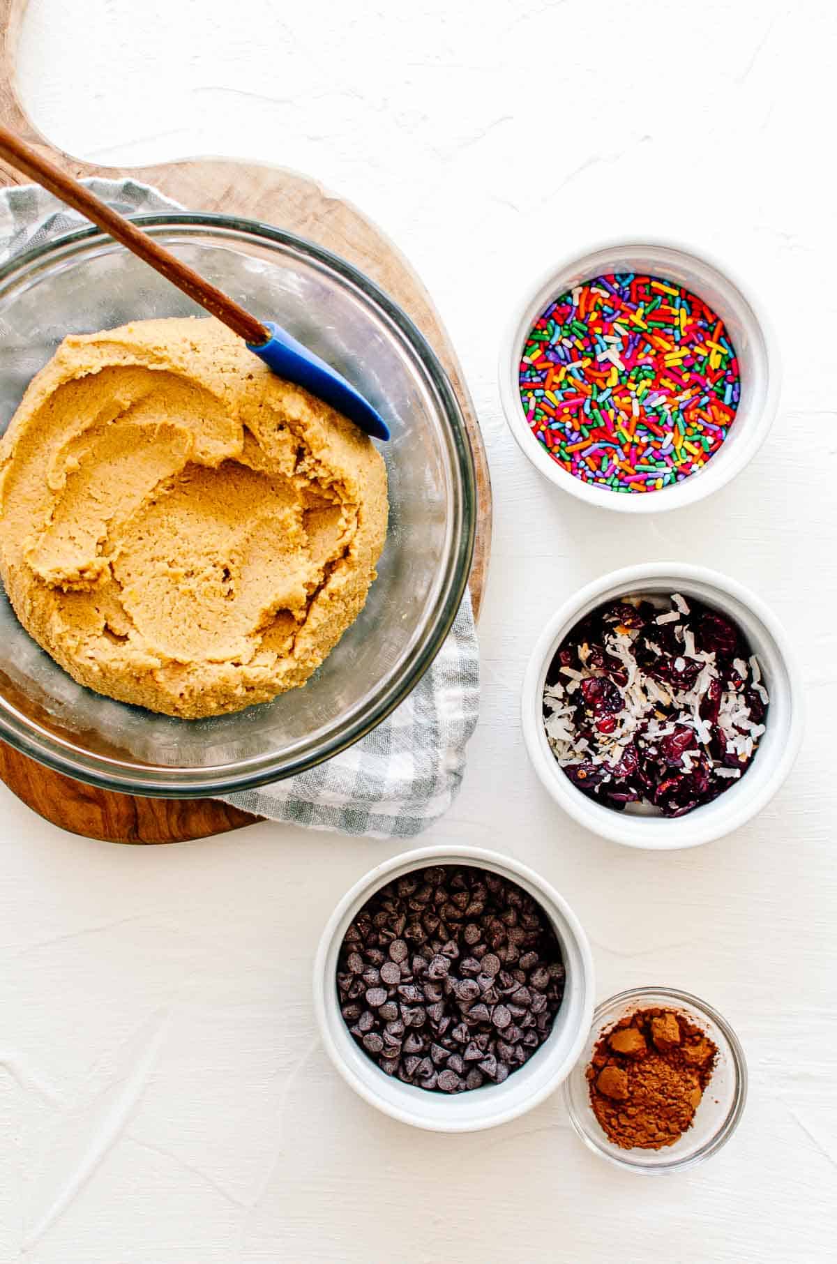 L'impasto per biscotti di ceci in una ciotola con diversi add ins porzionati sul lato, tra cui scaglie di cioccolato, cacao in polvere, mirtilli rossi e cocco, e sprinkles.